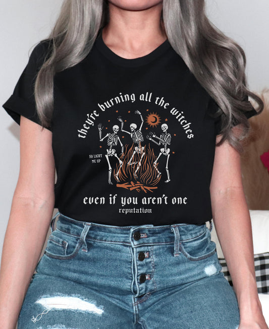 Woman wearing a black graphic t-shirt that says “They’re burning all the witches even if you aren’t one – Reputation,” featuring dancing skeletons around a fire, inspired by Taylor Swift’s Reputation era.