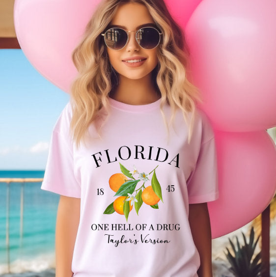 Woman wearing a pastel pink tee with oranges and "Florida: One Hell of a Drug – Taylor’s Version" printed on the front, holding pink balloons near a beach
