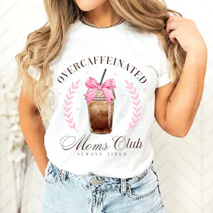 Woman wearing a white t-shirt that says “Overcaffeinated Moms Club – Always Tired” with a pink bow-topped iced coffee graphic, styled with light denim shorts.