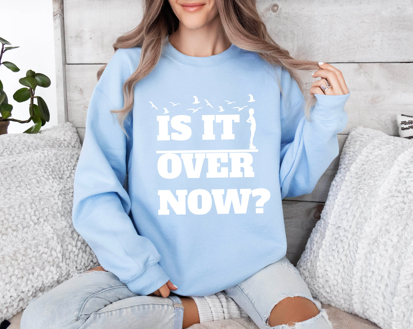 Woman wearing a light blue crewneck sweatshirt with the words “Is It Over Now?” in bold white text and bird silhouette graphic above.