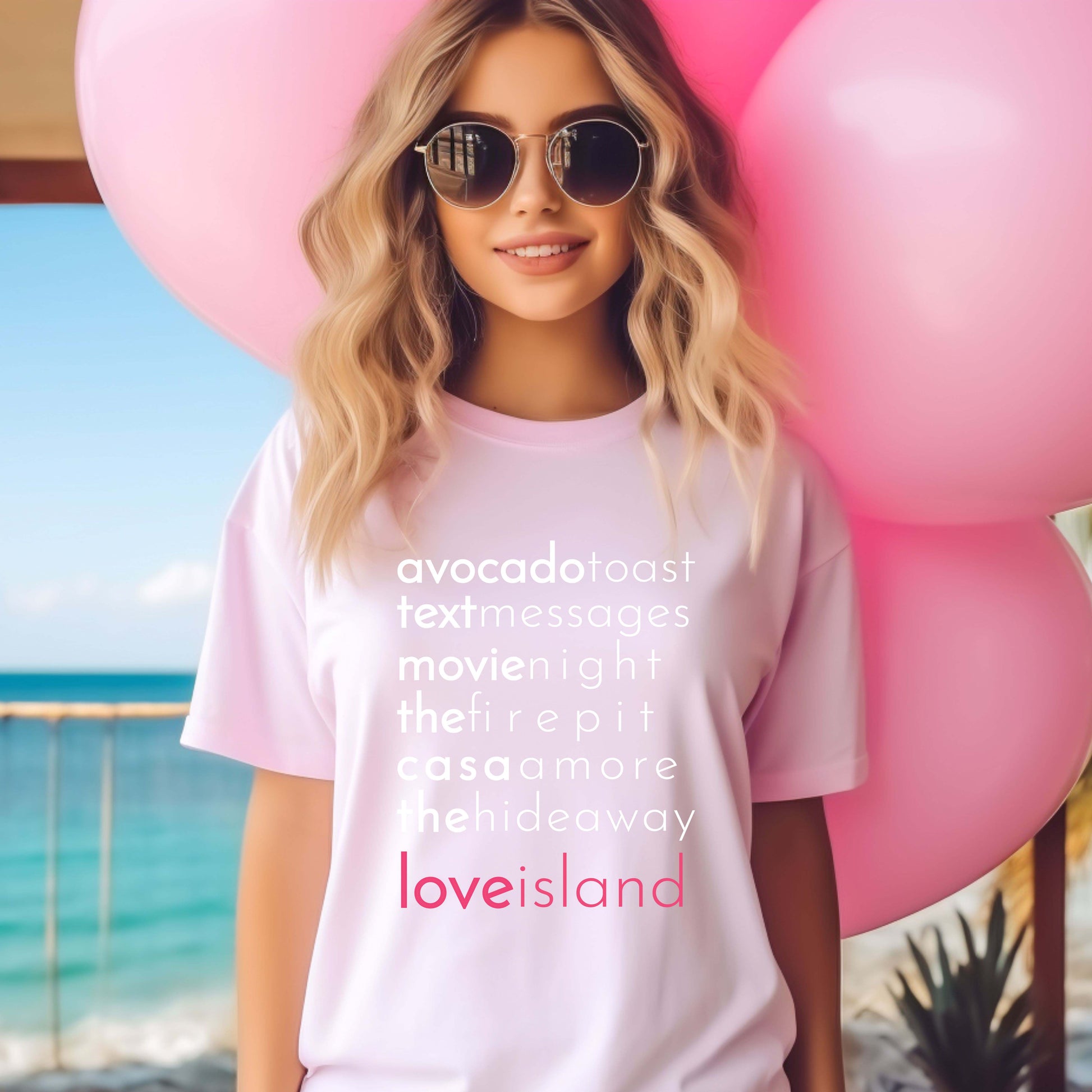 Woman wearing a pink t-shirt with Love Island-inspired phrases like “avocado toast,” “the fire pit,” and “casa amor,” holding pink balloons on a beachside deck.