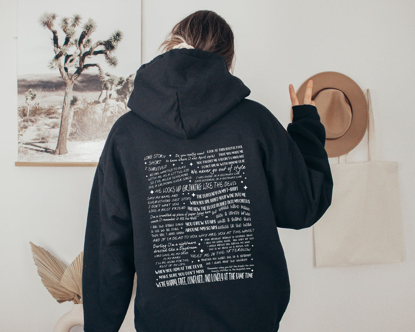 Person wearing a black hoodie with Taylor Swift quote collage design on the back, flashing a peace sign in a cozy neutral room