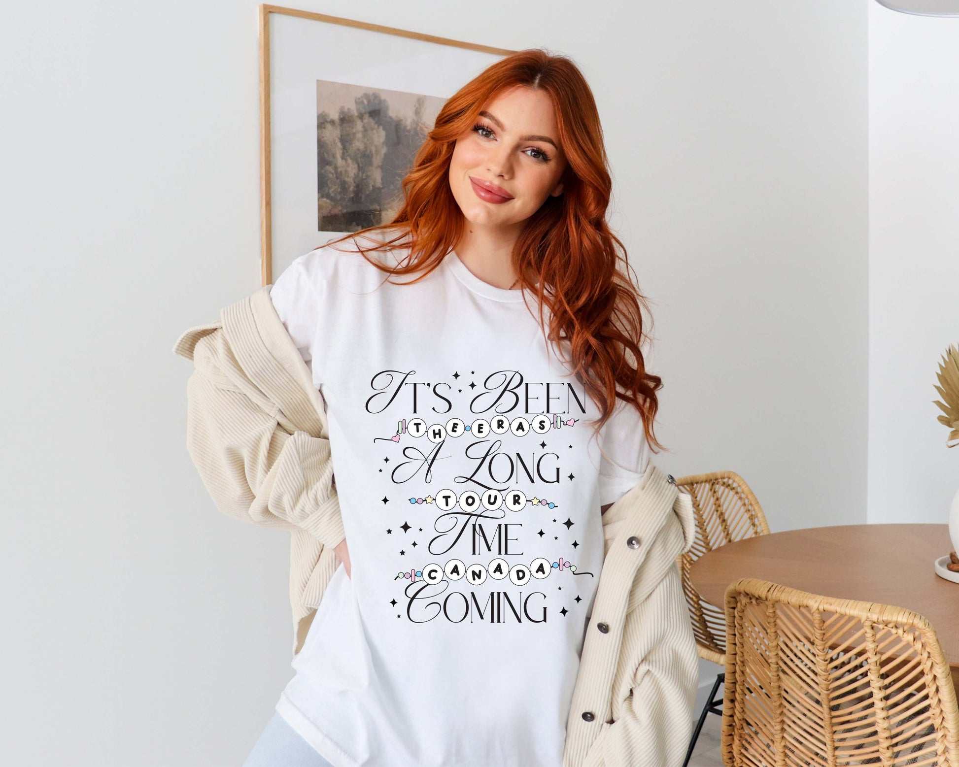 Red-haired woman wearing a white Taylor Swift-inspired t-shirt that reads “It’s Been a Long Time Coming” with The Eras Tour and Canada spelled out in friendship bracelet lettering
