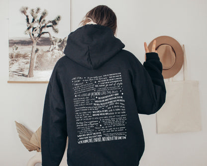 Person wearing a black hoodie with Taylor Swift quote collage design on the back, flashing a peace sign in a cozy neutral room